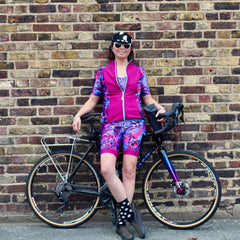 Rouleur Leggings View B - image of woman in brightly coloured cycling shorts and jersey standing in front of a bicycle and a brick wall
